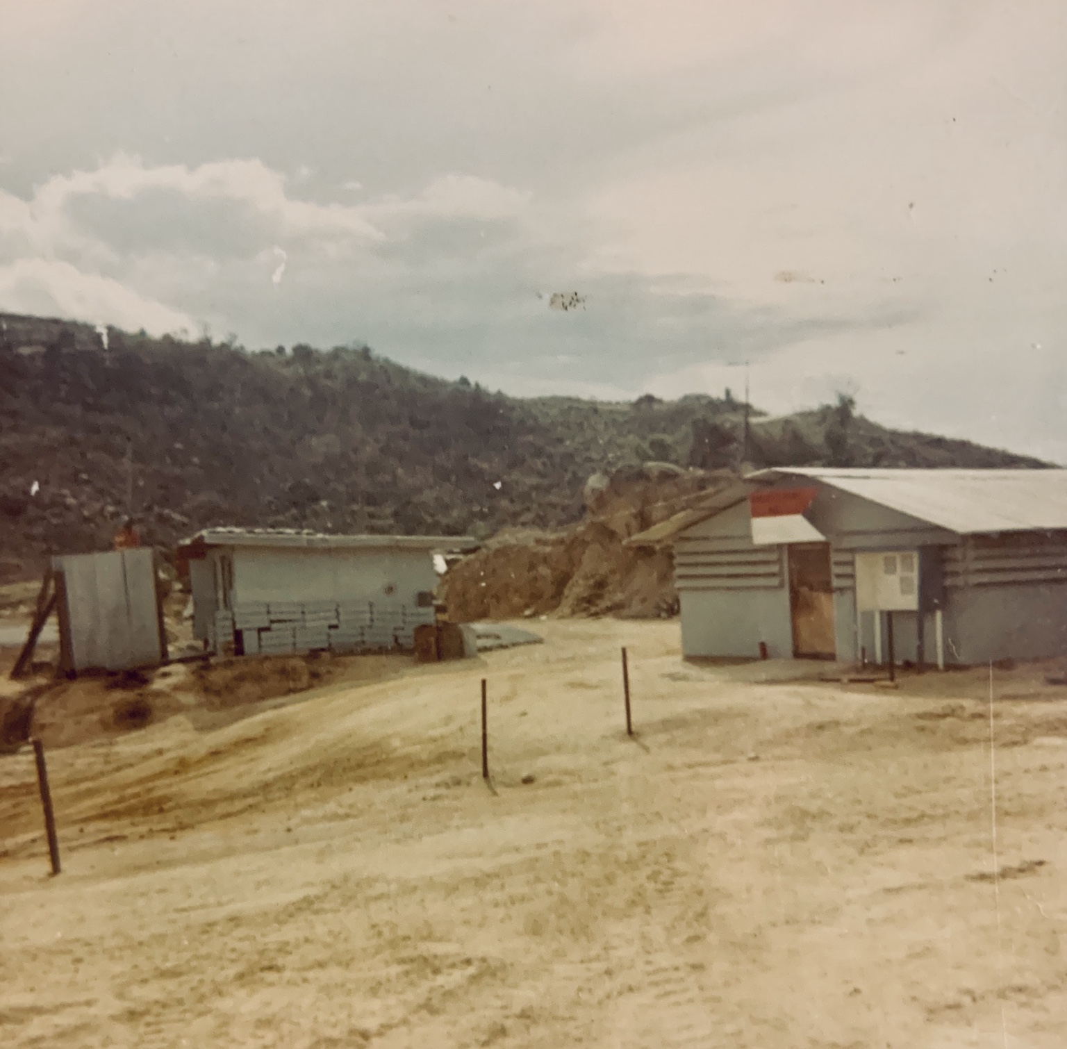 Base buildings and a dirt road below a wooded hillside, believed to be at LZ Bayonet.