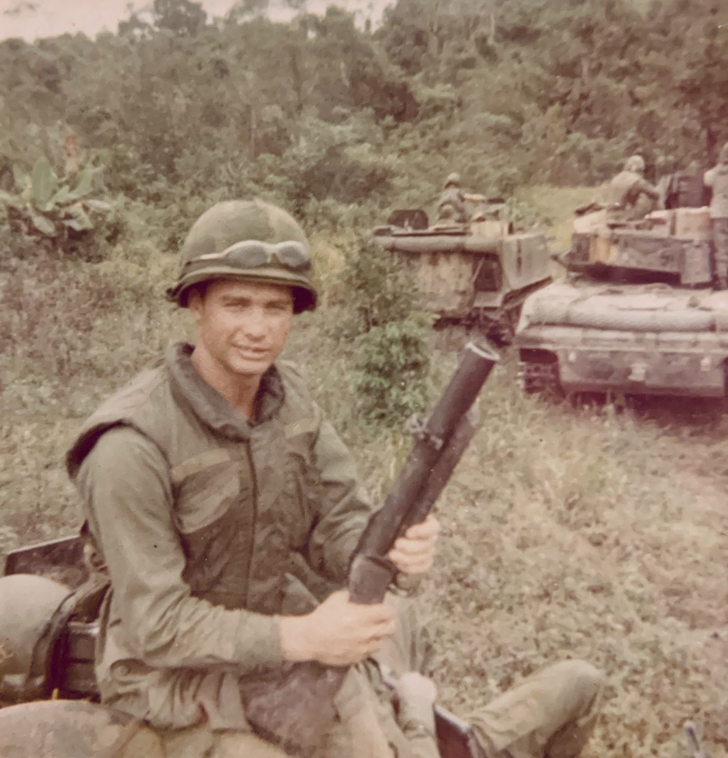 Stephen Rowley holding a shoulder-fired launcher, with H Troop tracks and troopers behind him in dense terrain.