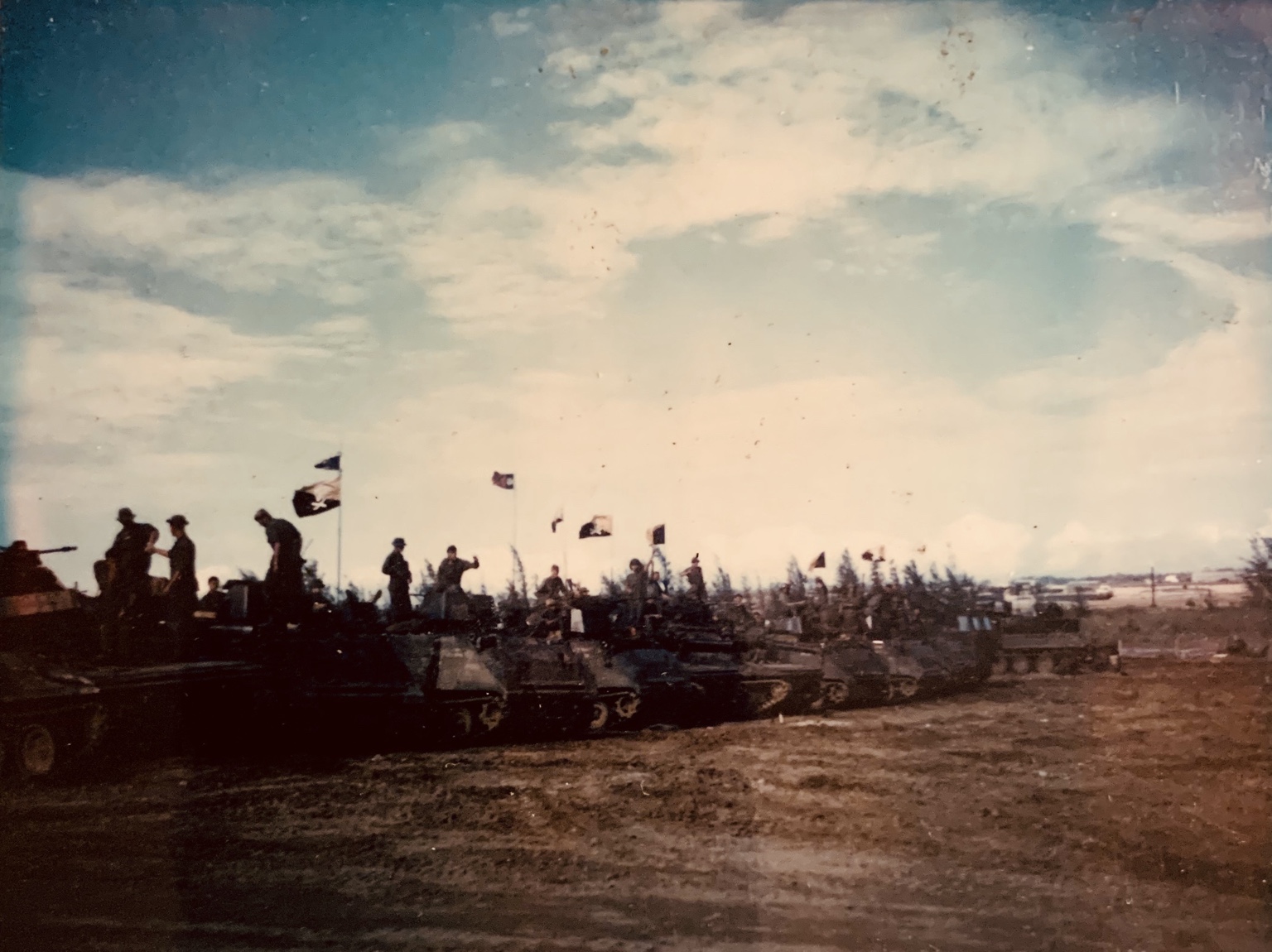 A long formation of H Troop tracks and their crews displaying flags in an open field.