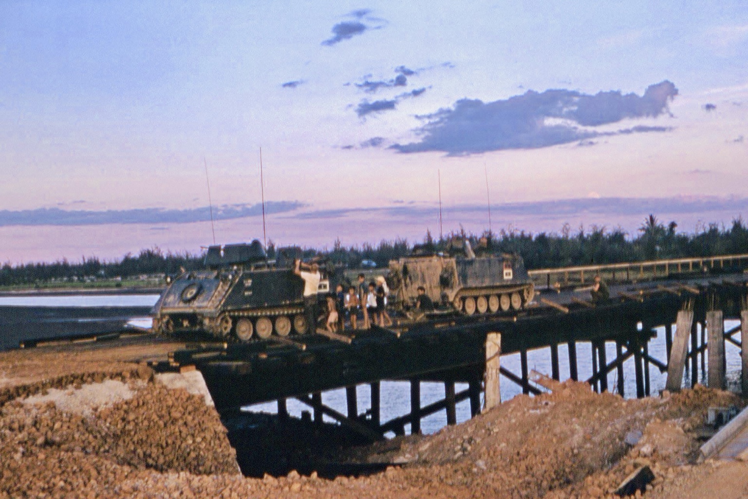 Two armored tracked vehicles on a bridge over water, with several people gathered nearby beneath a pink-and-blue sky, 1968. Bridge and individuals unidentified.