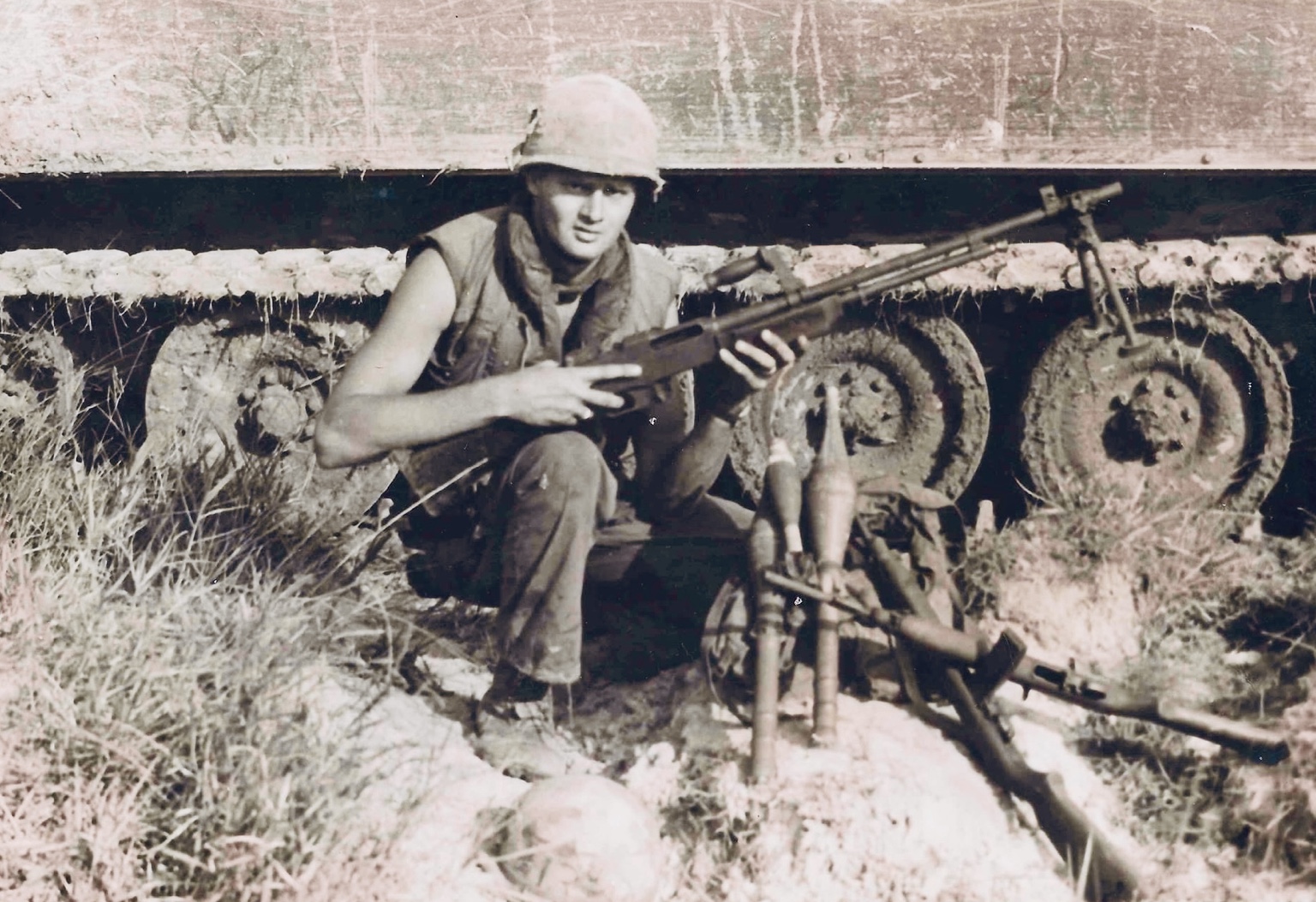 Trooper Charles Watson kneeling beside an armored tracked vehicle, wearing a helmet and flak vest and holding a weapon. Date and location unknown.