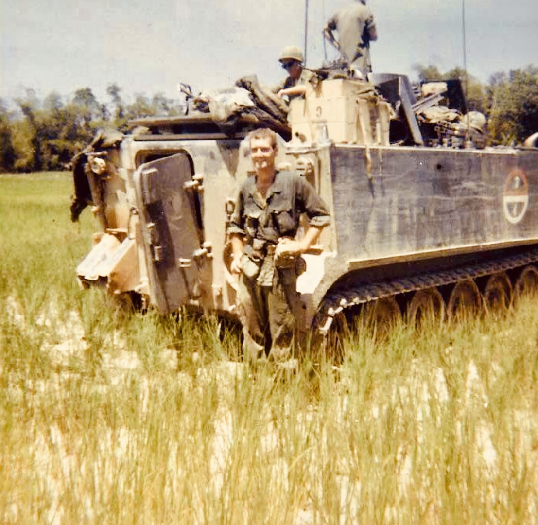 An unidentified trooper beside an armored tracked vehicle in a grassy field, with fellow troopers aboard. Date and location unknown.