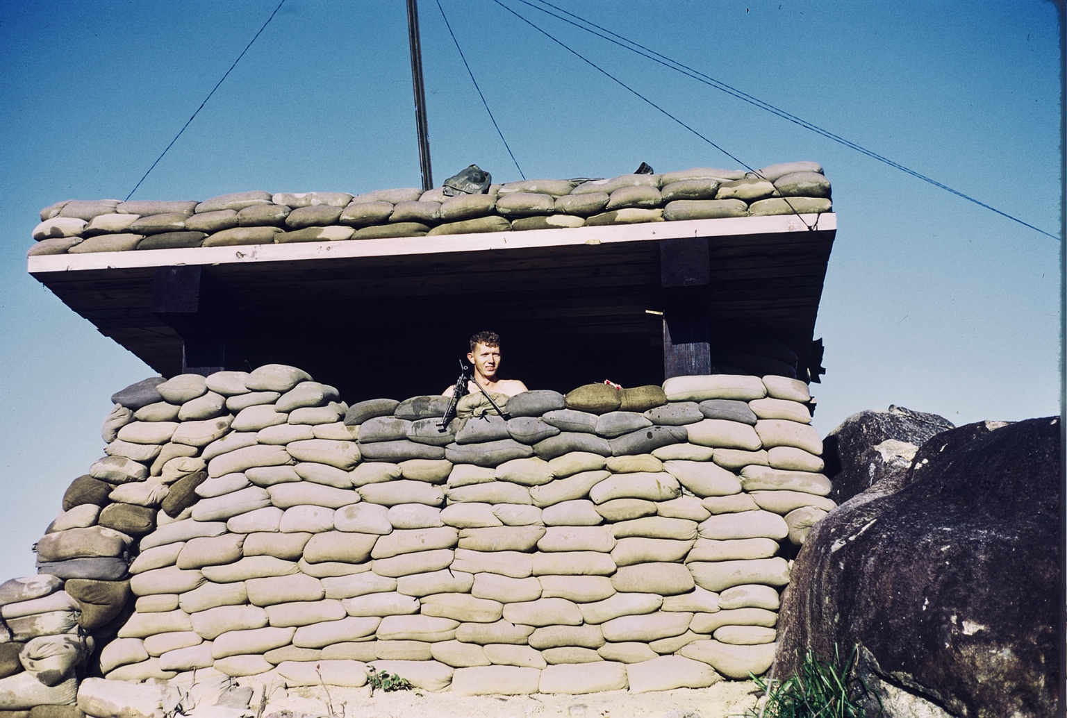 An unidentified H Trooper with a rifle standing behind a heavily sandbagged defensive position.