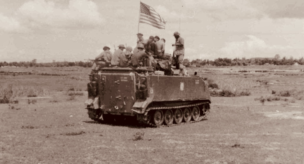 H Troopers riding atop an M113 armored personnel carrier flying the American flag in an open field in Vietnam.